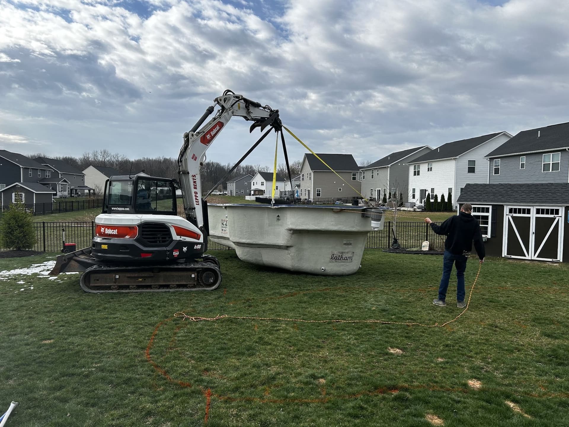 Compact excavator with lifting straps attached to a white fiberglass pool shell in a fenced backyard, while a worker stands nearby holding a line.