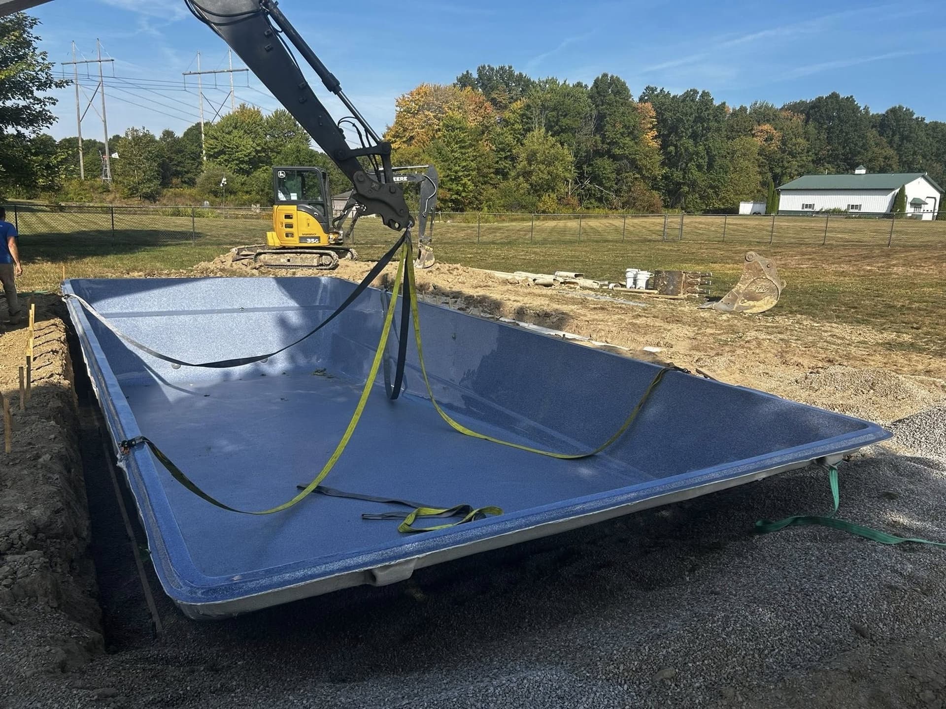 Excavator arm setting a large rectangular fiberglass pool shell onto a gravel base in an open rural lot, with a second excavator and a barn in the background.