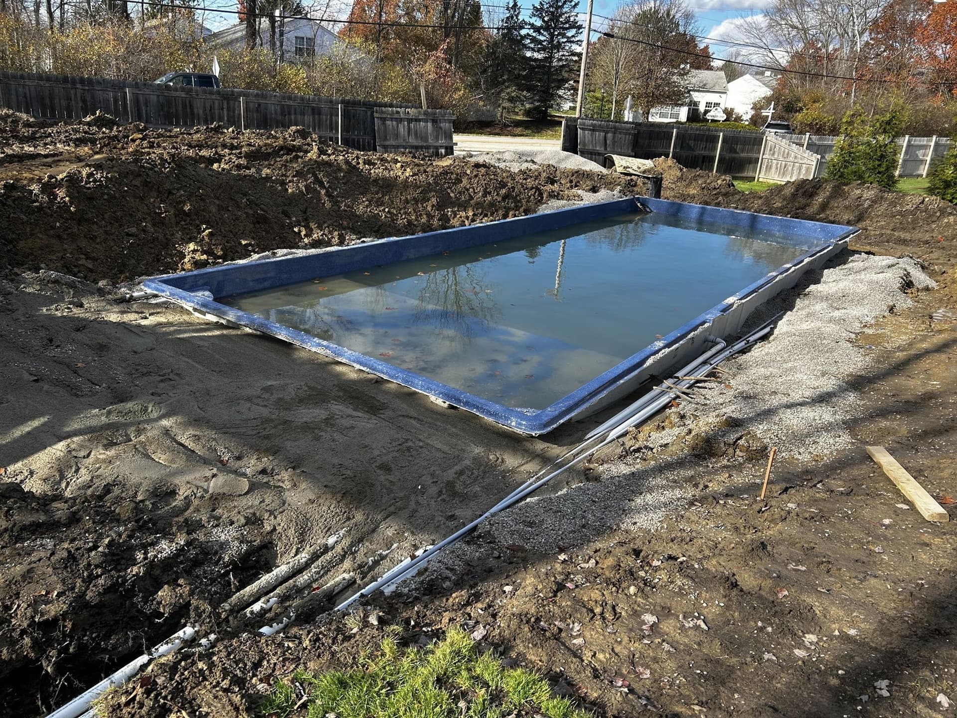 Rectangular fiberglass pool with a blue interior partly filled with water, set in a fresh excavation with plumbing lines running along one side.