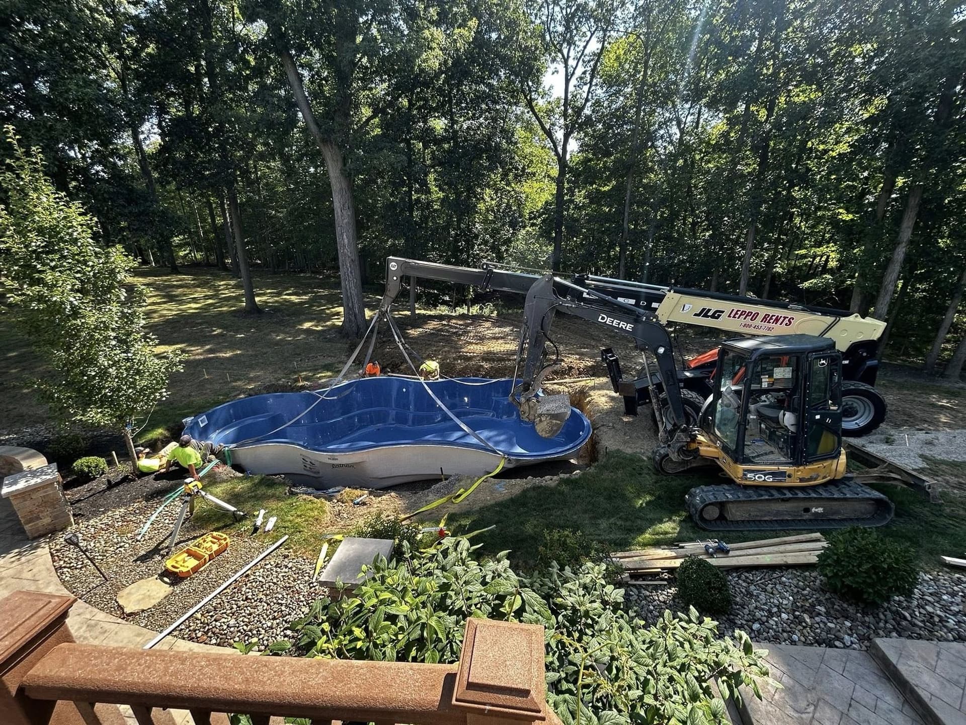 View from a backyard deck of a freeform fiberglass pool shell being set into a wooded lot, with an excavator, crew members and landscaping around the work area.