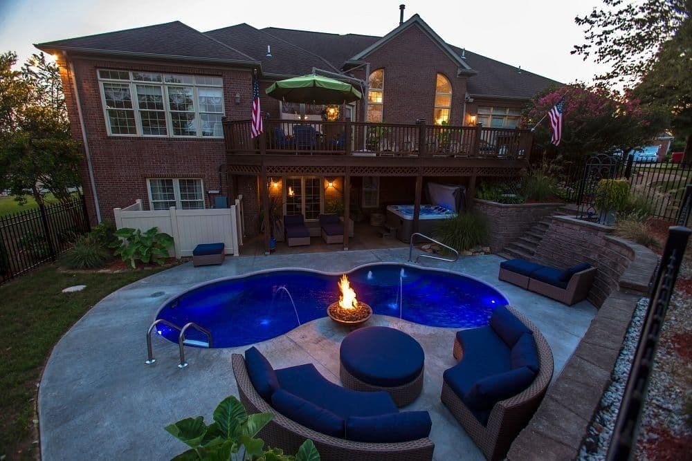 Freeform pool lit blue at dusk with a central fire pit and curved outdoor sofas on a concrete patio below a brick house with a deck.