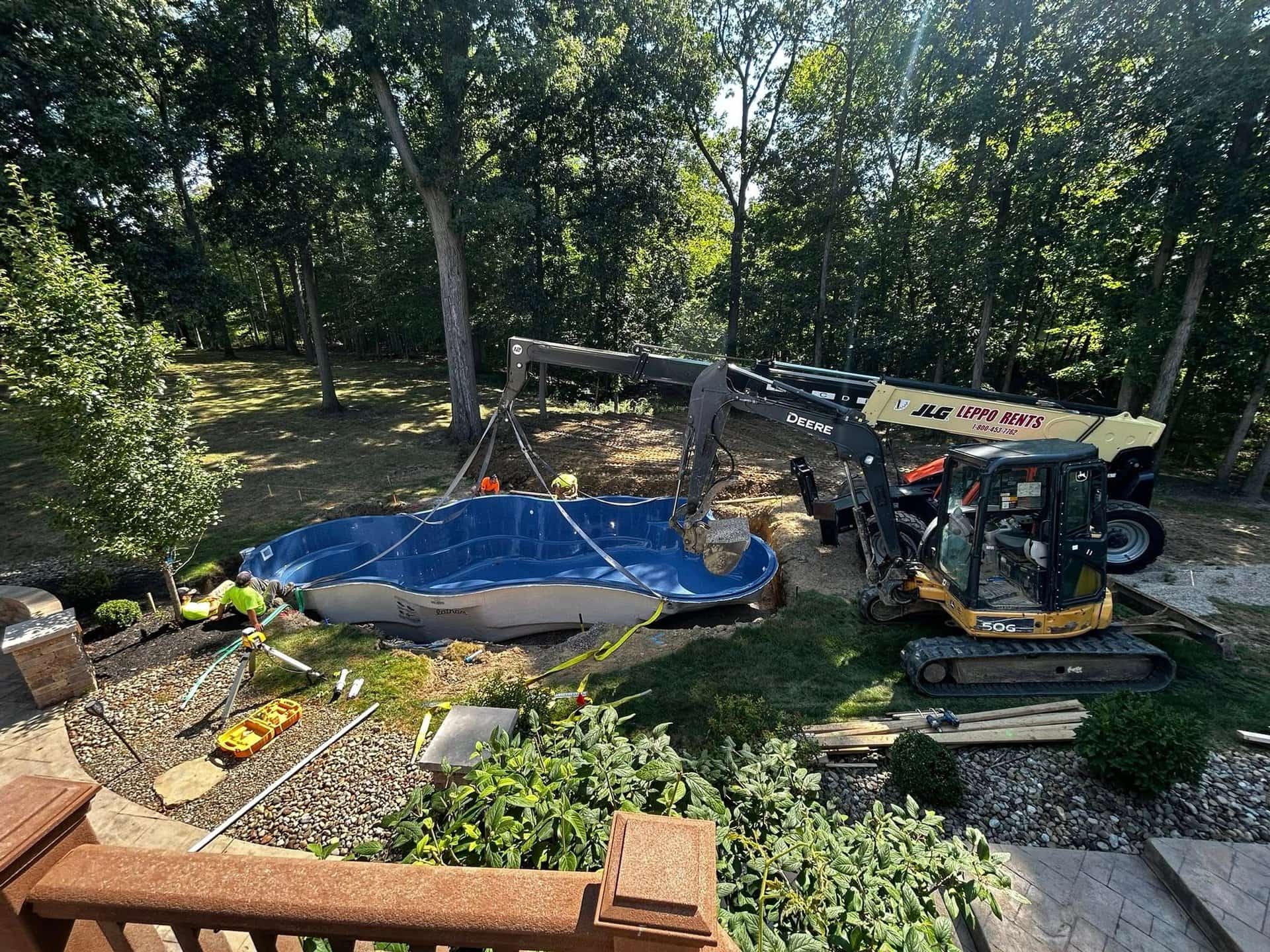Ohio State Pools crew setting a blue fiberglass pool shell with a crane and excavator in a wooded Northeast Ohio backyard
