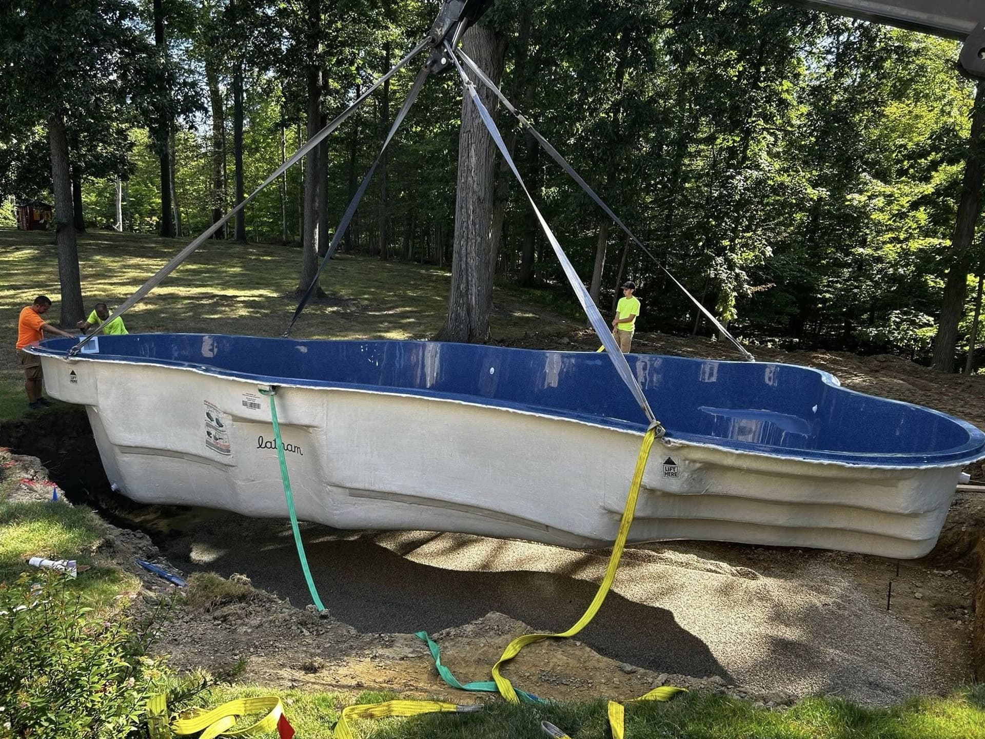 Freeform fiberglass pool shell with a blue interior hanging from lifting straps over a prepared gravel base in a wooded backyard, with two workers in safety shirts nearby.