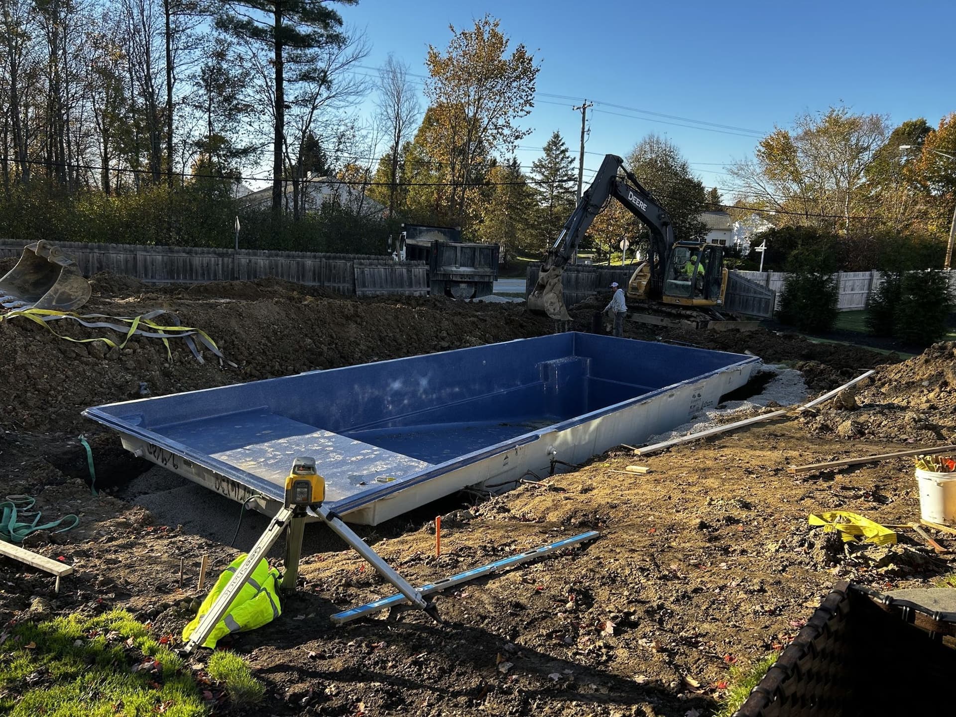 Blue rectangular fiberglass pool shell sitting in a freshly excavated backyard, with a laser level on a tripod in the foreground and an excavator working behind it.