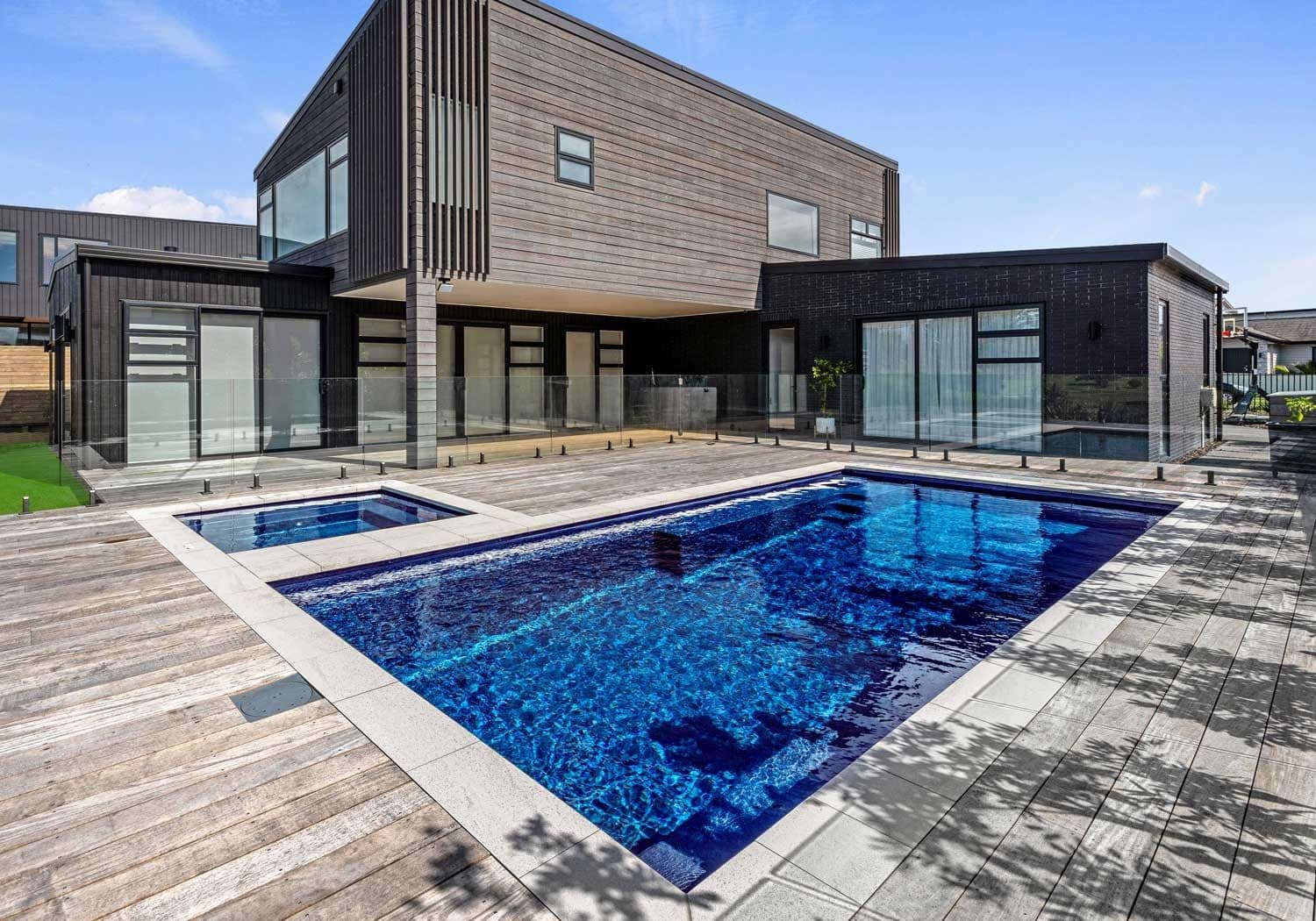 Liberty Compassion fiberglass pool, rectangular pool with a deep blue interior and an attached spa, a gray wood deck and a modern two-story house, manufacturer photo.