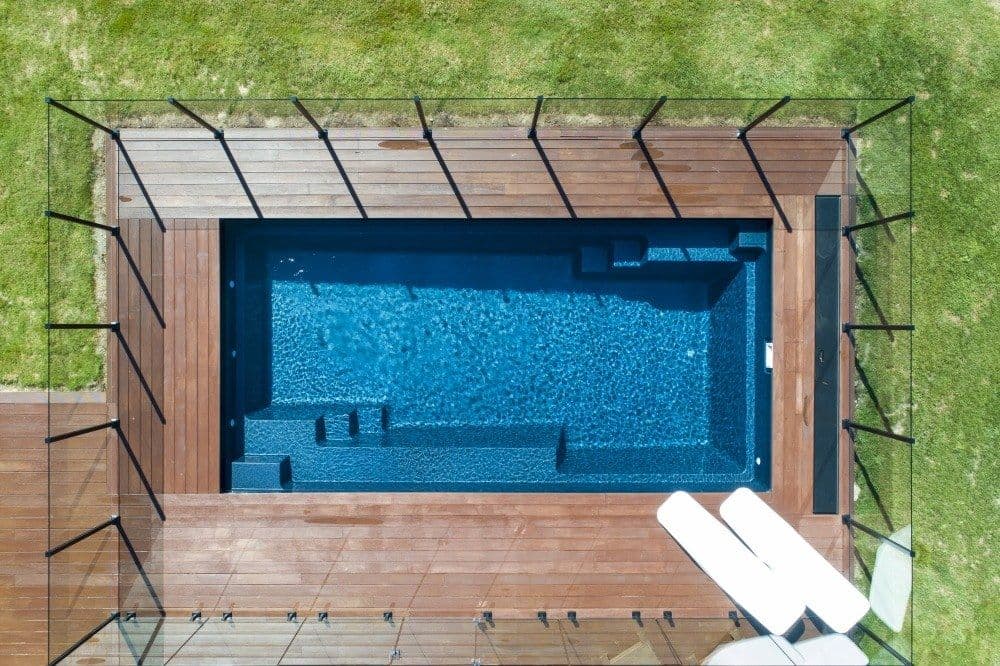 Liberty Compassion fiberglass pool, overhead view of a small rectangular pool with entry steps, framed by a wood deck, with two white loungers at one corner, manufacturer photo.