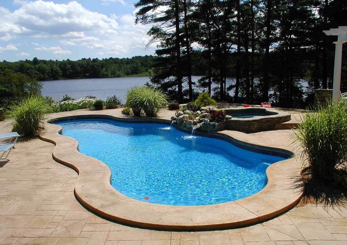 Liberty Devotion fiberglass pool, freeform pool with a tan concrete deck and a rock feature, next to a lake lined with trees, manufacturer photo.