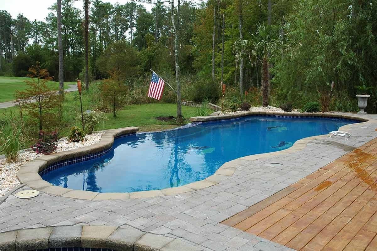 Liberty Devotion fiberglass pool, freeform pool with a stone border and a wood deck section, surrounded by trees and a lawn with a flagpole, manufacturer photo.
