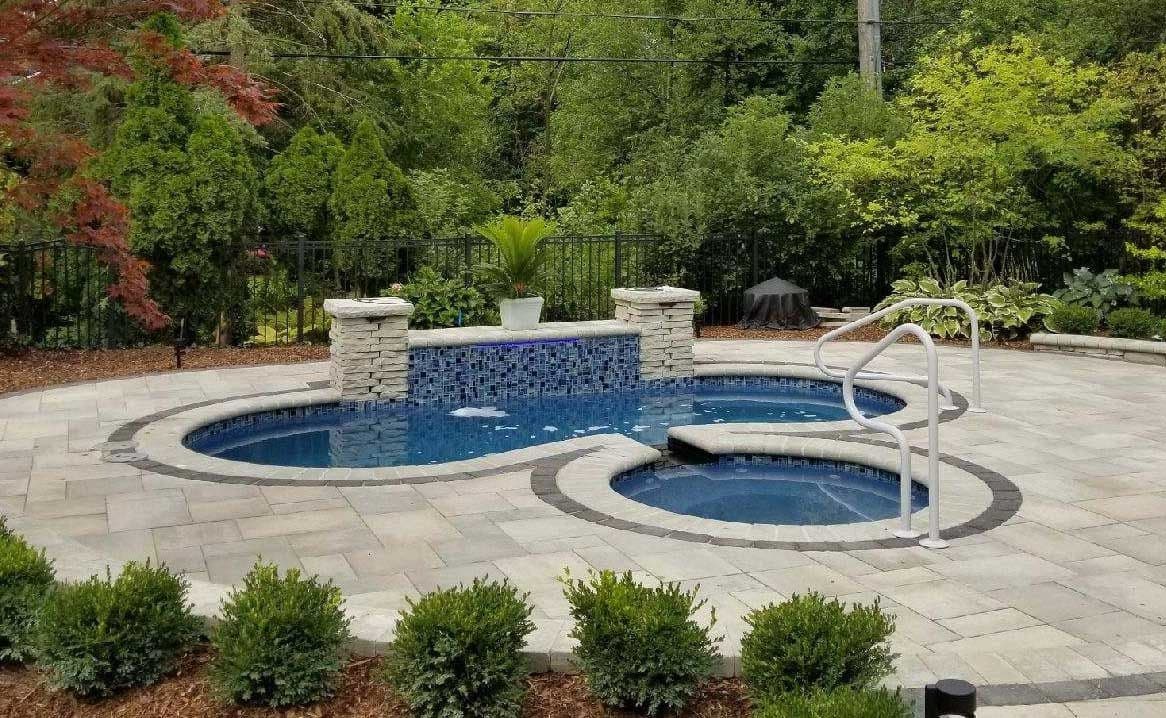Liberty Dignity fiberglass pool, kidney-shaped pool with a separate round spa and a stone water wall, set in a gray paver patio with shrubs, manufacturer photo.