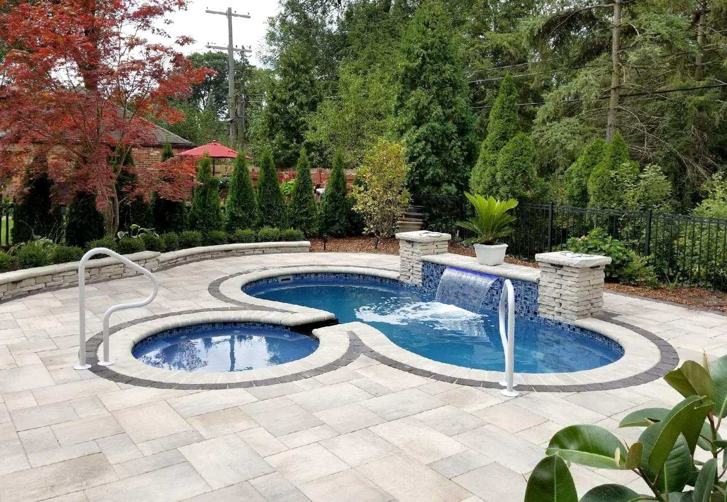 Liberty Dignity fiberglass pool, kidney-shaped pool and round spa with a water spillway, gray pavers and a stone pillar wall in a wooded yard, manufacturer photo.