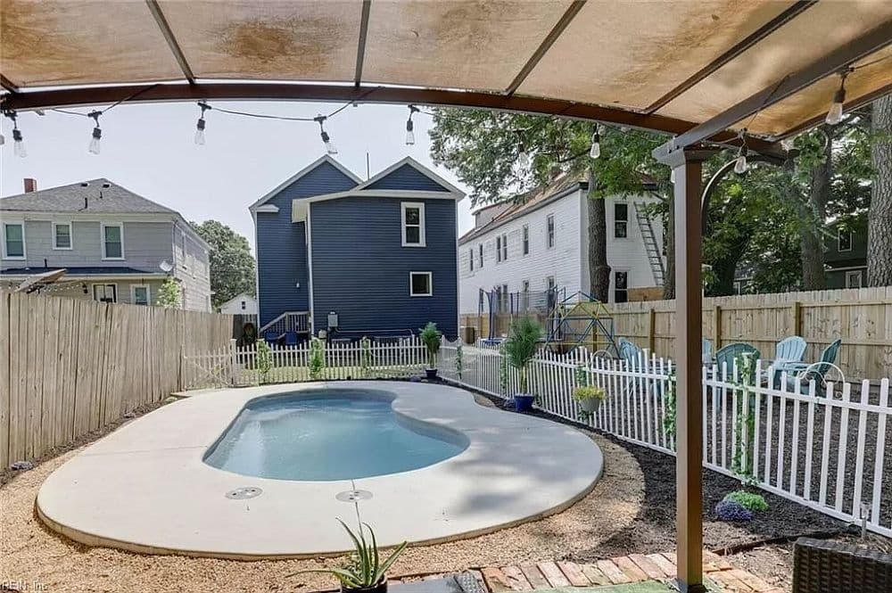 Liberty Dignity fiberglass pool, small kidney-shaped pool with a light concrete deck under a pergola, beside a white picket fence and a house, manufacturer photo.