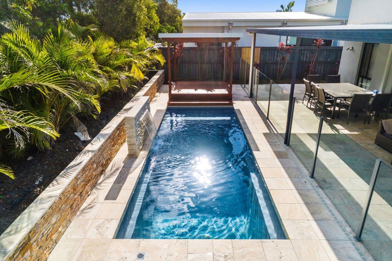 Liberty Enrichment fiberglass pool, long narrow rectangular pool with a stone-clad wall, glass fence and a covered outdoor room at the far end, manufacturer photo.