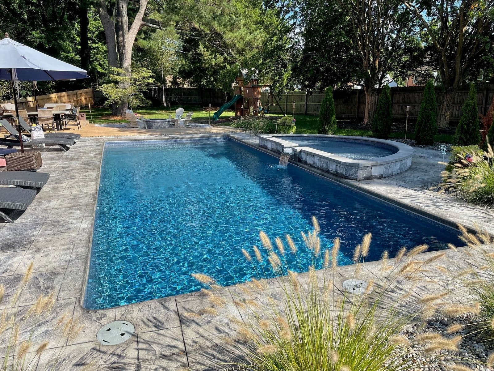 Liberty Glory fiberglass pool, long rectangular pool with a blue tile interior, gray stone deck and an attached raised spa in a tree-lined yard, manufacturer photo.