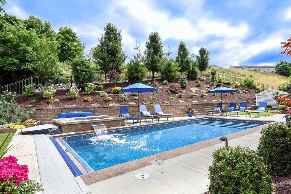 Liberty Glory fiberglass pool, rectangular pool with a stone-edged terrace, blue umbrellas, lounge chairs and a flowering hillside, manufacturer photo.