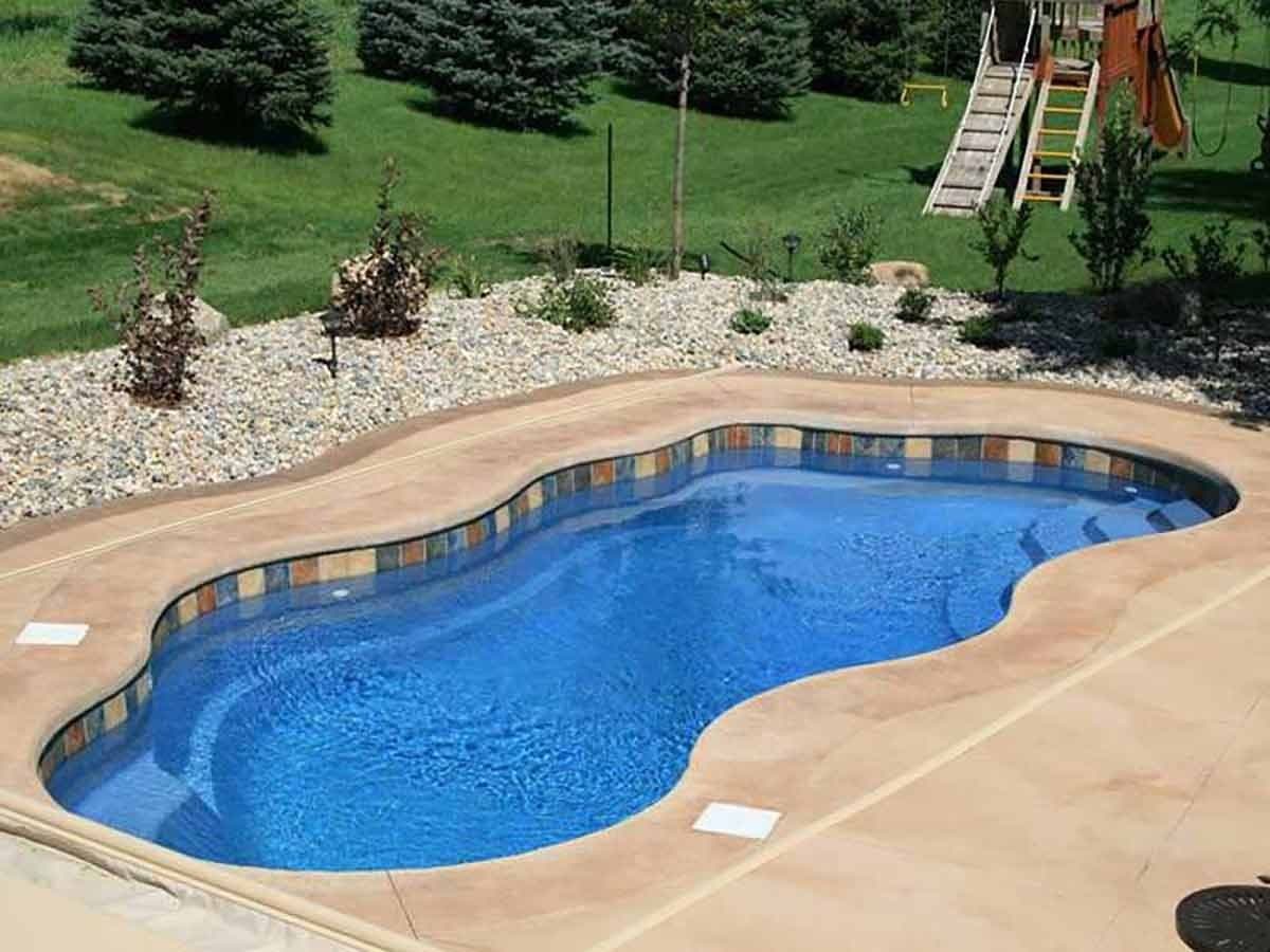 Liberty Hero fiberglass pool, freeform pool with a blue interior and curved tan concrete deck, in a yard with evergreens and a swing set, manufacturer photo.