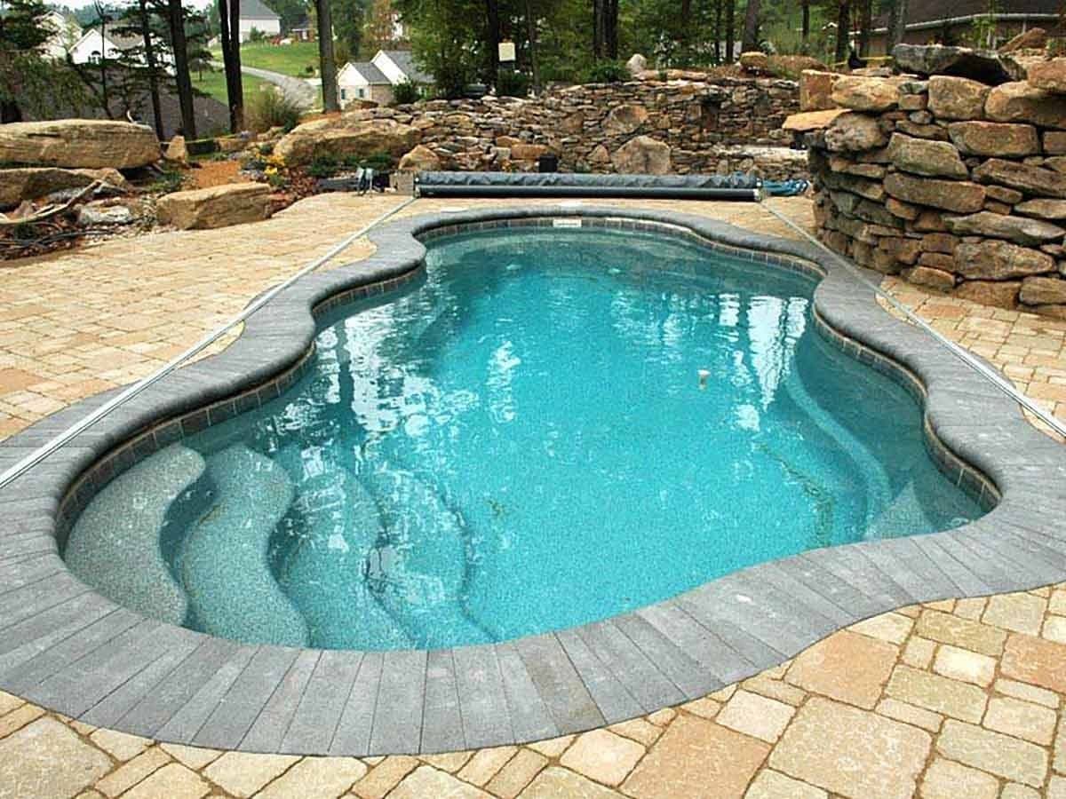 Liberty Hero fiberglass pool, freeform pool with entry steps, stone coping and a paver deck in front of a stacked stone wall, manufacturer photo.