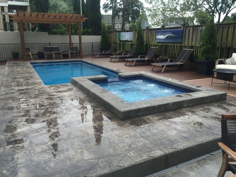 Liberty Independence fiberglass pool, rectangular pool with an attached raised spa on a gray stone patio, with a pergola and lounge chairs, manufacturer photo.