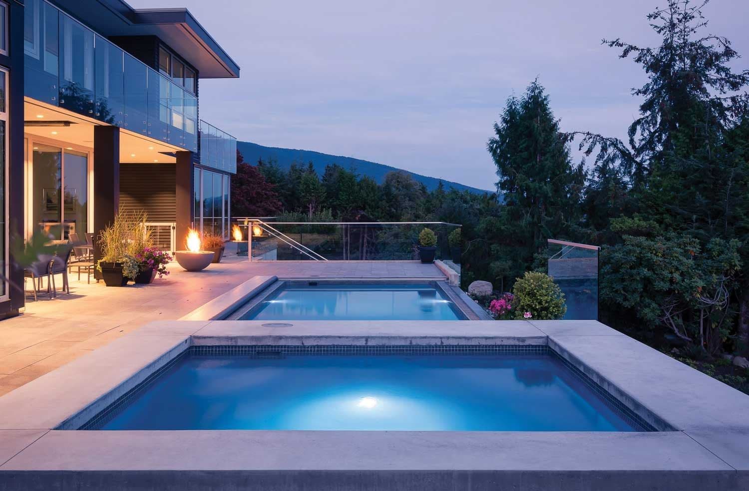 Liberty Independence fiberglass pool, rectangular pool and attached spa lit at dusk beside a modern glass-walled house, manufacturer photo.