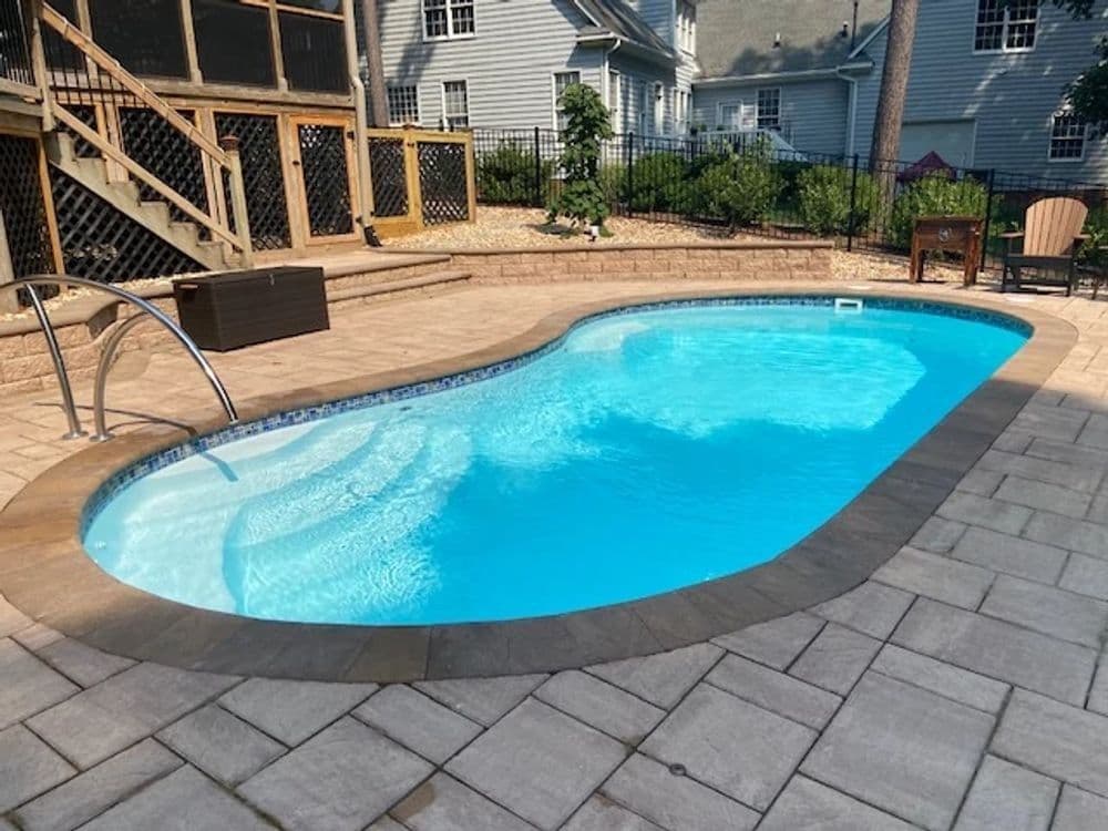 Liberty Merit fiberglass pool, kidney-shaped pool with a light blue interior and gray paver deck, with a handrail and a house behind, manufacturer photo.