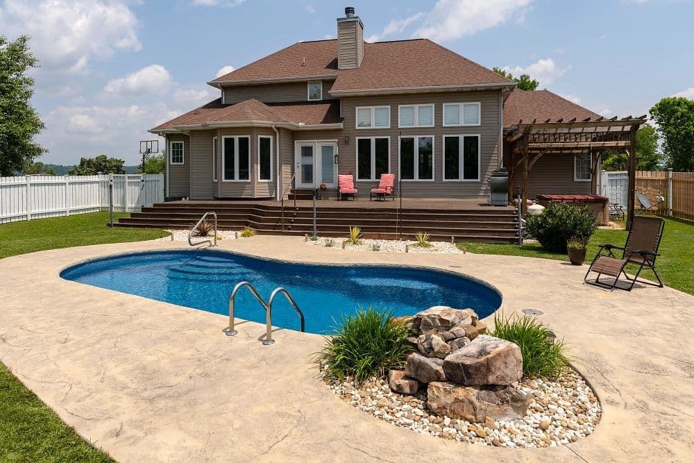 Liberty Merit fiberglass pool, kidney-shaped pool with a concrete deck and a rock planter, in front of a brick house with a pergola, manufacturer photo.