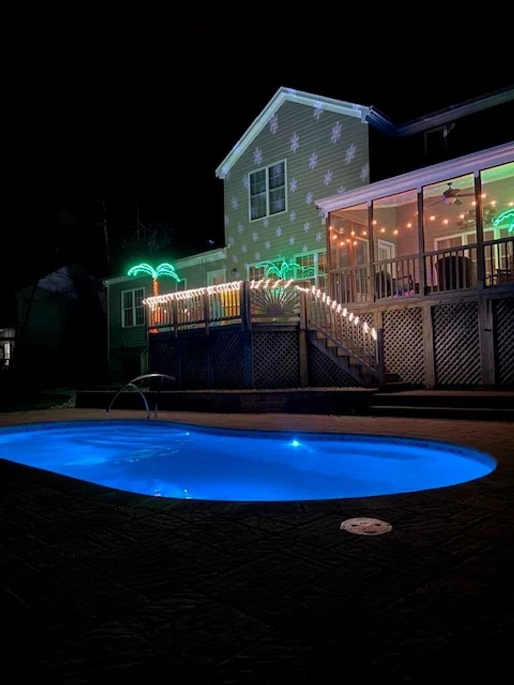 Liberty Merit fiberglass pool, kidney-shaped pool lit blue at night below a house deck with string lights, manufacturer photo.