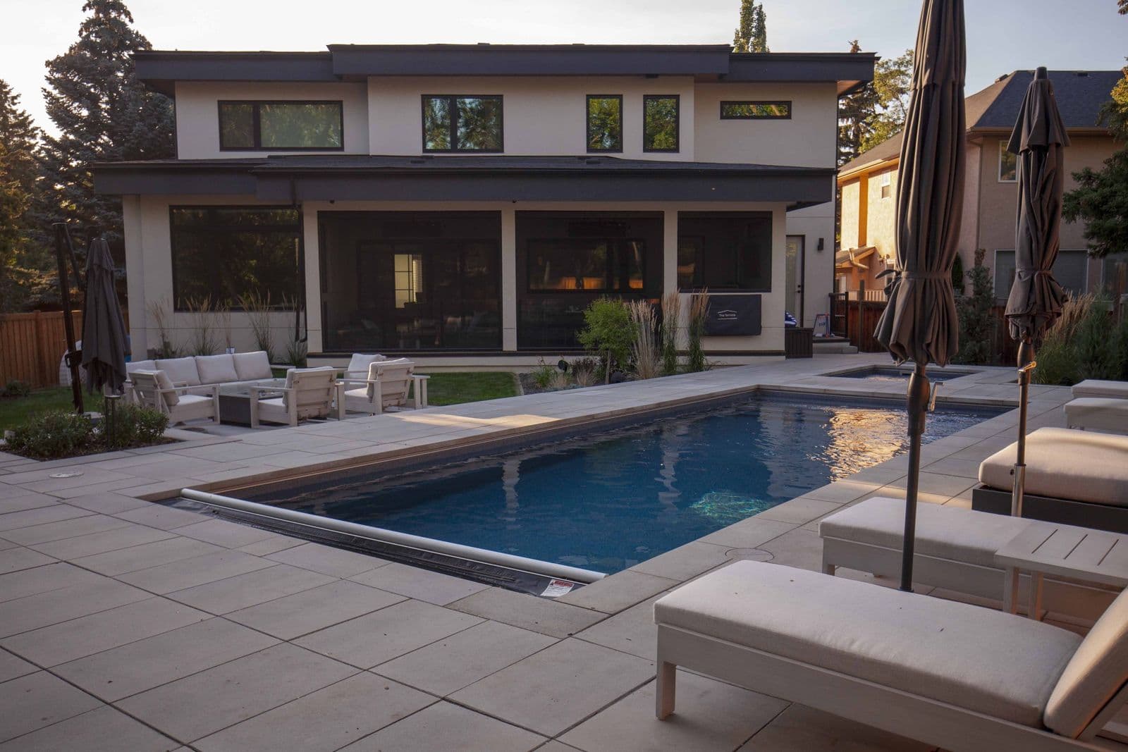 Liberty Opportunity fiberglass pool, rectangular pool with a dark interior, stone deck, lounge chairs and umbrellas beside a modern house at dusk, manufacturer photo.