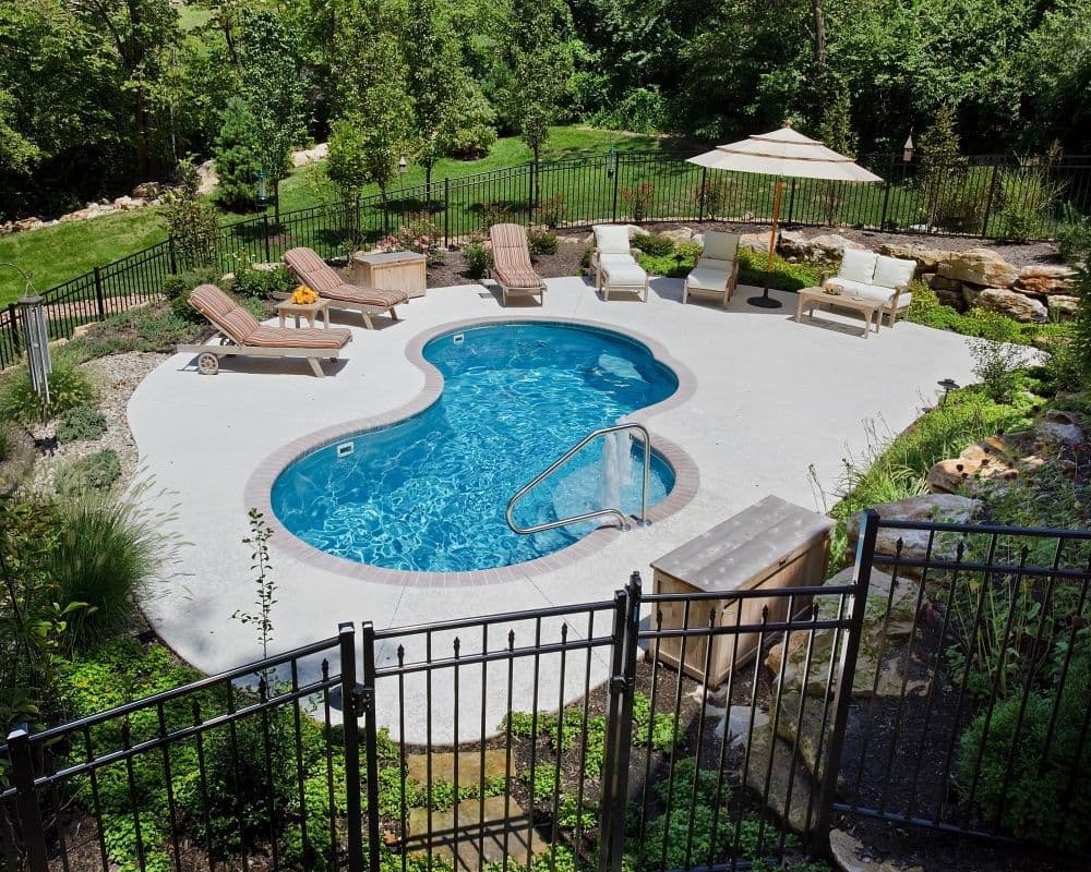 Liberty Poise fiberglass pool, freeform pool with a light concrete deck and lounge chairs, seen from above behind a black metal fence, manufacturer photo.