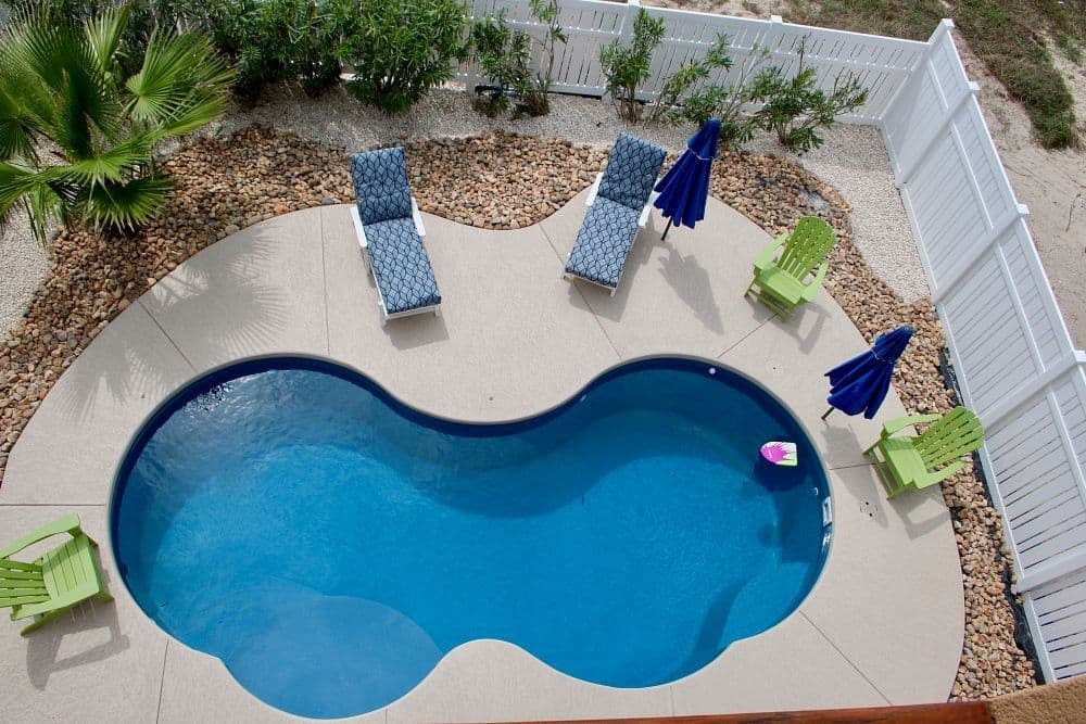 Liberty Poise fiberglass pool, freeform pool with a light concrete deck, blue lounge chairs and a white vinyl fence, seen from above, manufacturer photo.