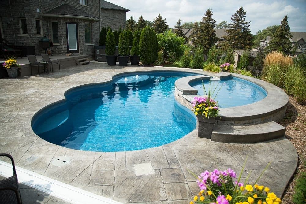 Liberty Poise fiberglass pool, freeform pool with a raised round spa, stamped concrete deck and flower beds, manufacturer photo.