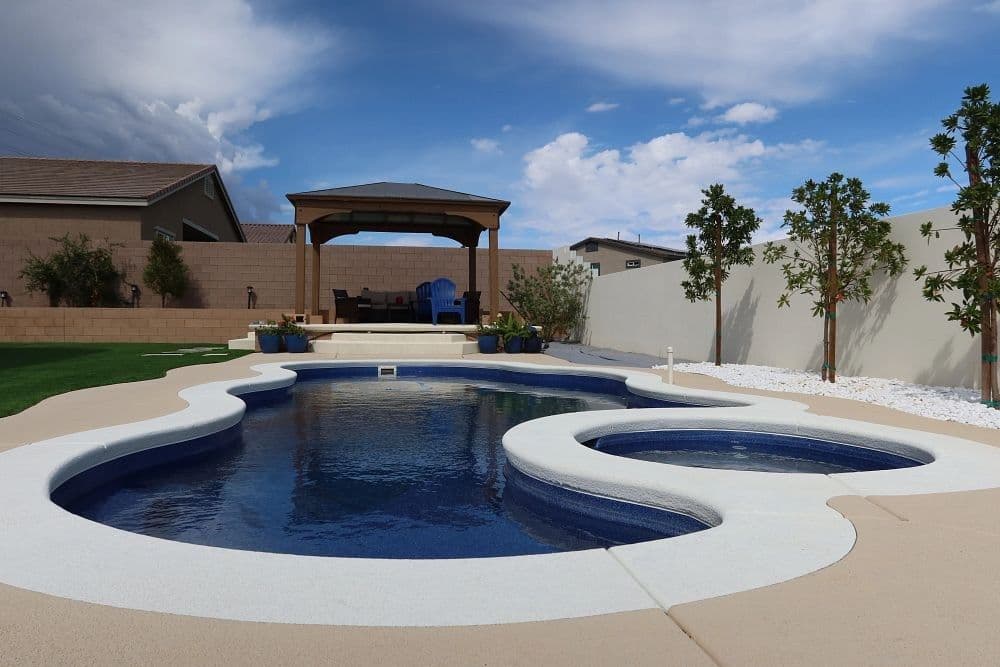Liberty Relaxation fiberglass pool, freeform pool with an attached round spa, a light concrete deck and a pavilion behind a white wall, manufacturer photo.