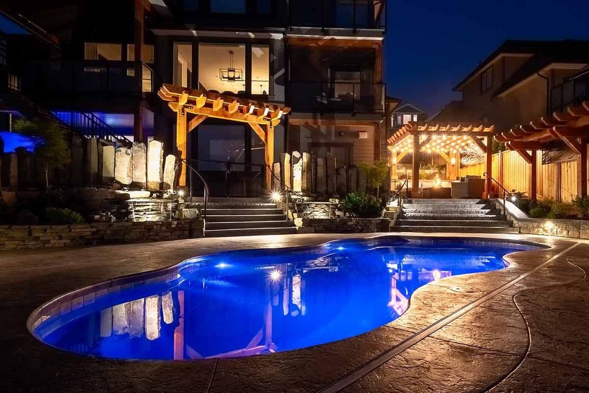 Liberty Relaxation fiberglass pool, freeform pool lit blue at night beside a lit pergola and patio, manufacturer photo.