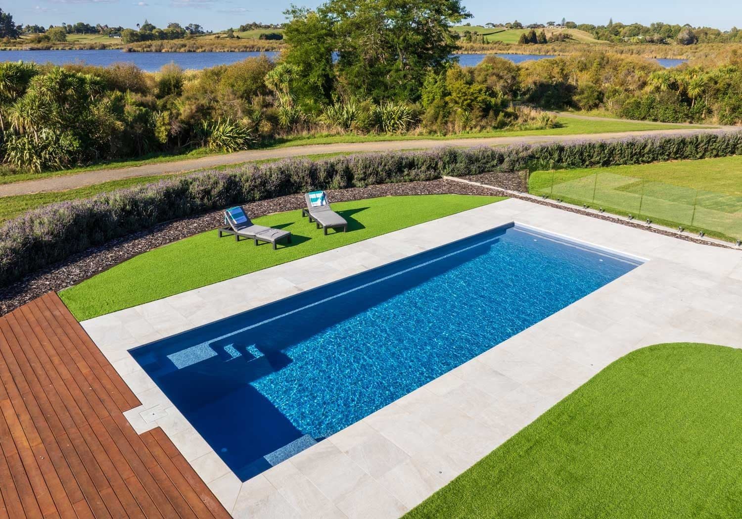Liberty Stature fiberglass pool, long rectangular pool with entry steps, a wood deck and lawn, with a pond and fields in the background, manufacturer photo.