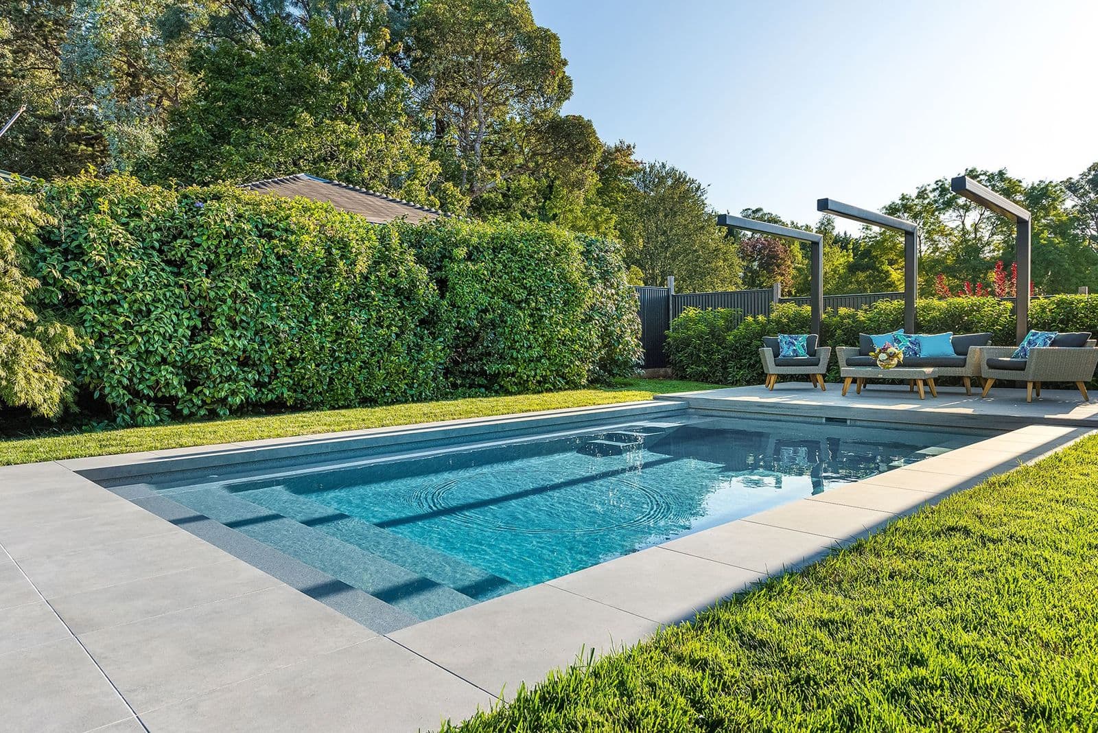Liberty Stature fiberglass pool, rectangular pool with wide entry steps, light stone deck and a hedge with a pergola behind, manufacturer photo.