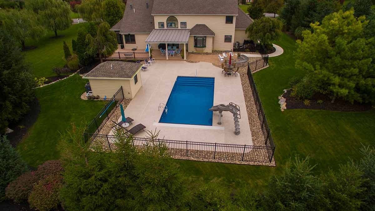 Liberty Tradition fiberglass pool, aerial view of a rectangular pool with a wide concrete deck behind a house, bordered by trees, manufacturer photo.