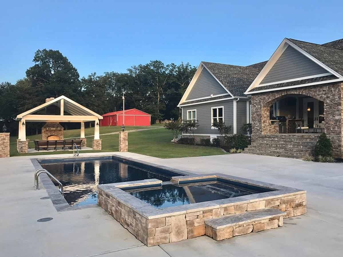 Liberty Tradition fiberglass pool, rectangular pool with an attached raised spa faced in stone, in front of a gray house and covered patio, manufacturer photo.