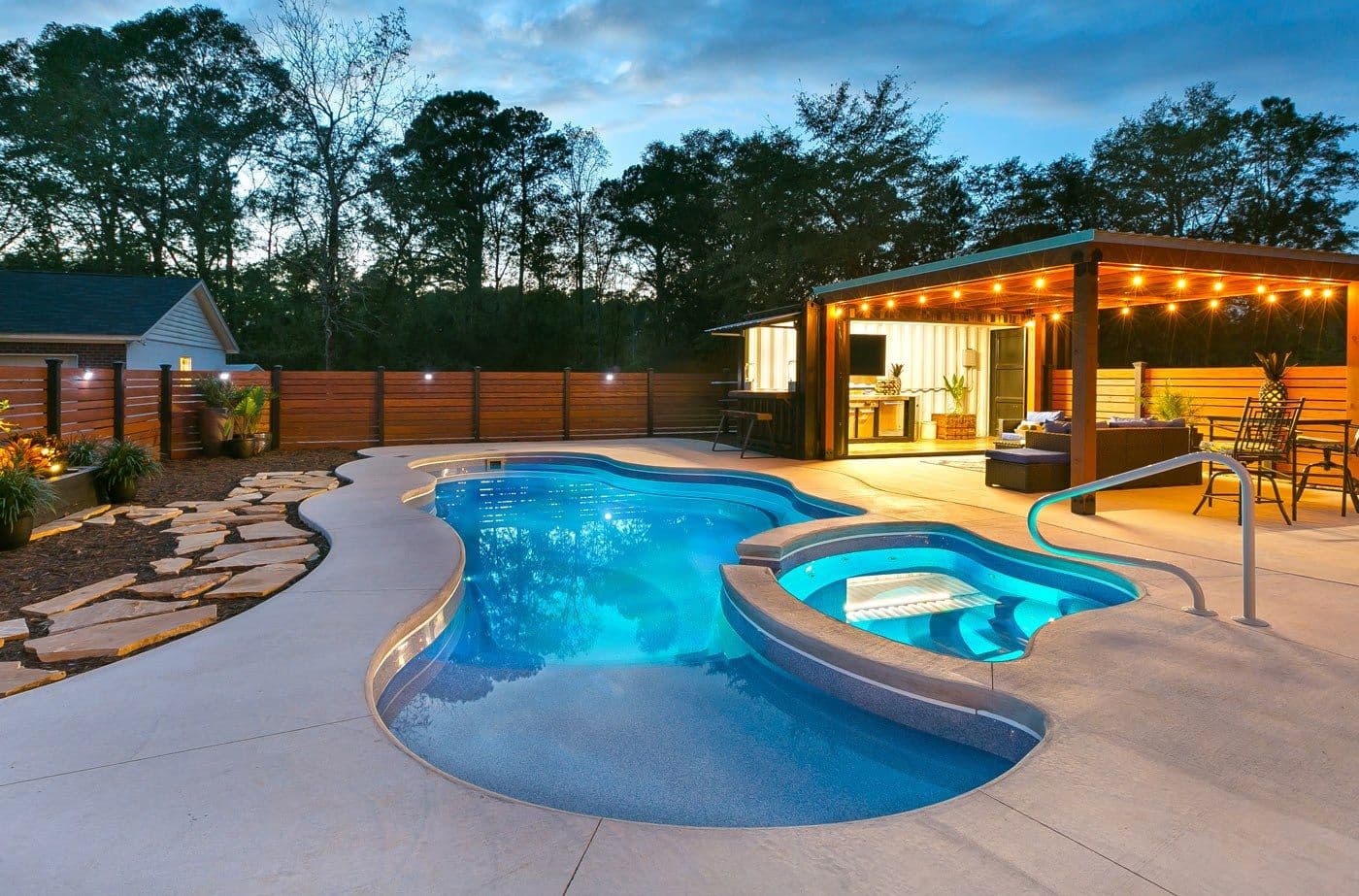 Liberty Unity fiberglass pool, freeform pool with an attached spa, stone deck and an outdoor kitchen bar lit at dusk, manufacturer photo.