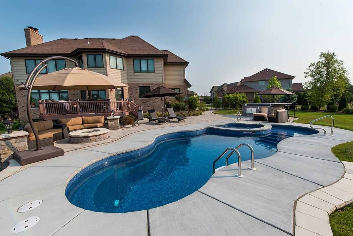 Liberty Unity fiberglass pool, freeform pool with a light concrete deck, a handrail and lounge area in front of a large house, manufacturer photo.