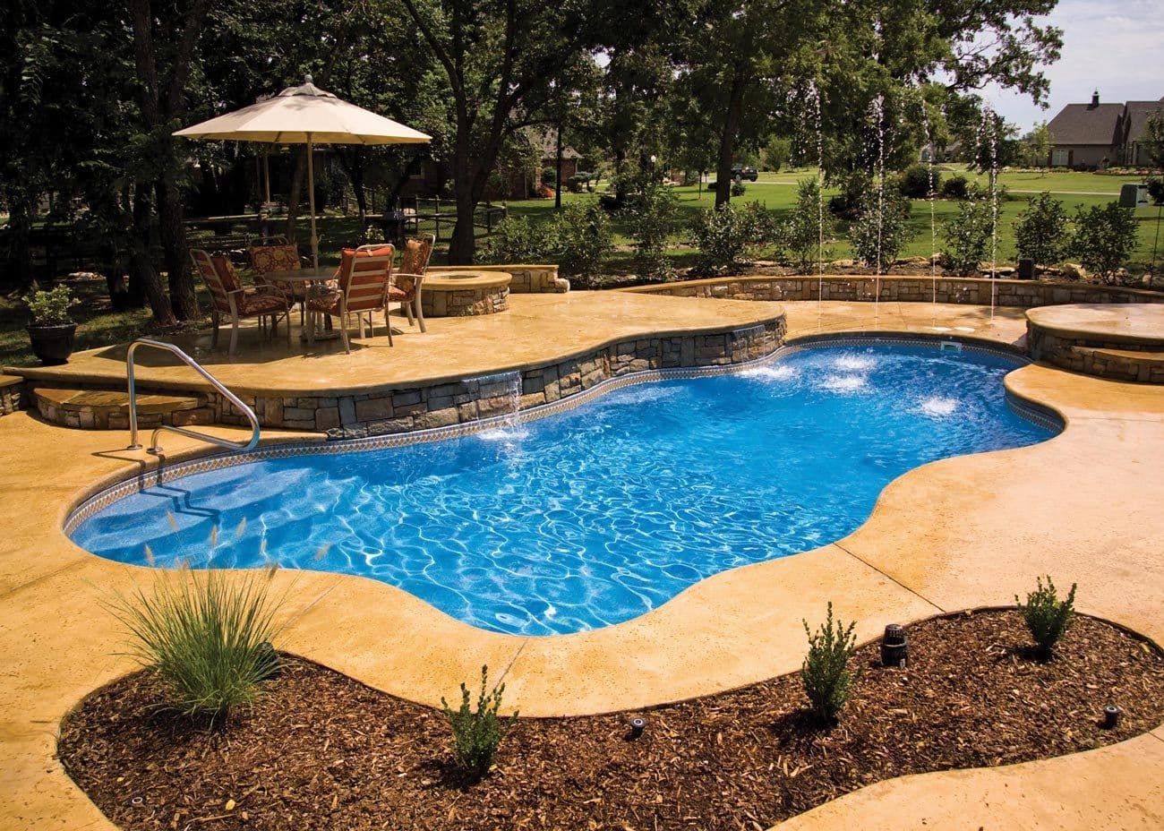 Liberty Unity fiberglass pool, freeform pool with a stamped concrete deck and landscaped border under a large tree, manufacturer photo.