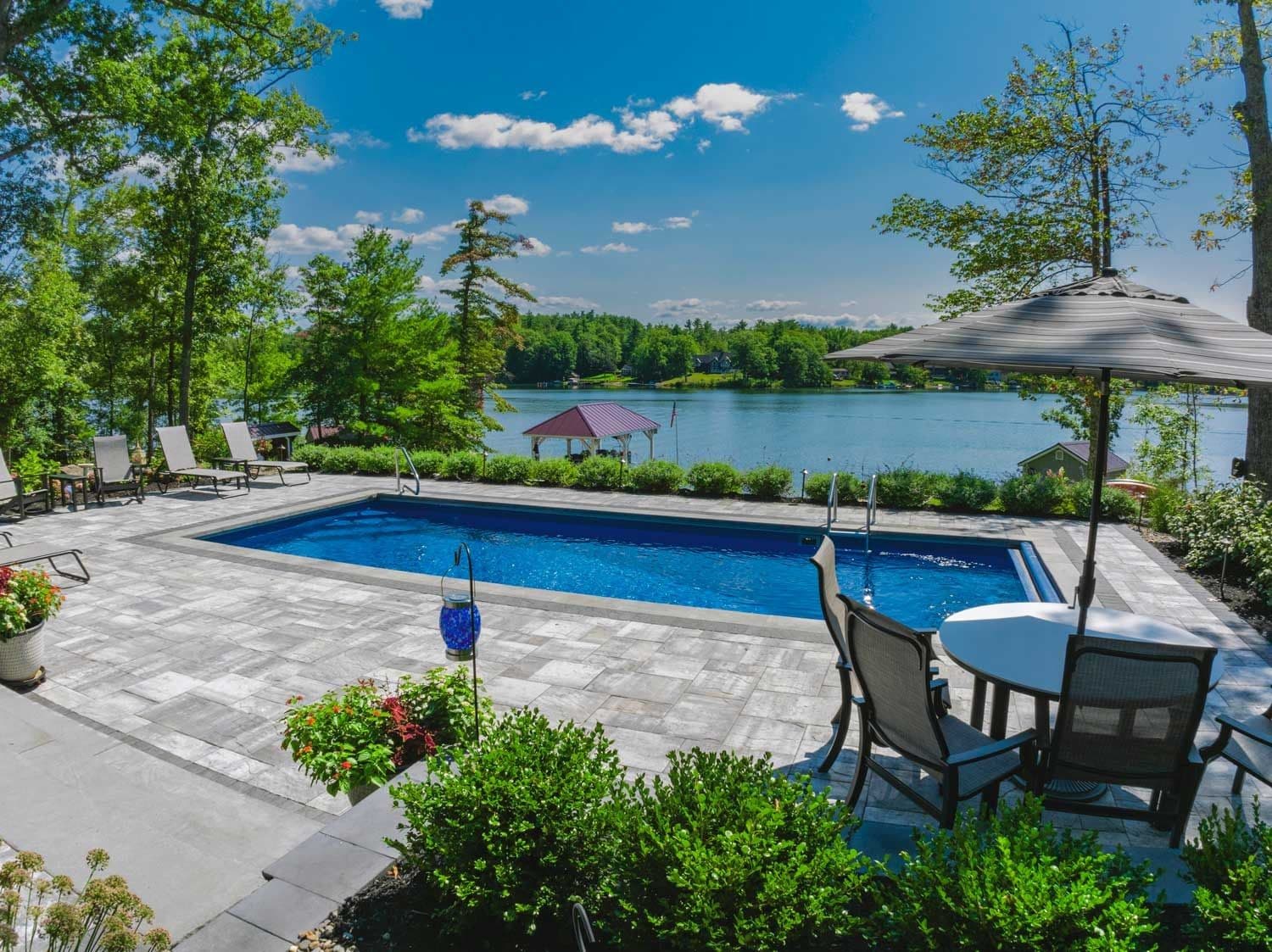 Liberty Valiant fiberglass pool, rectangular pool with gray stone deck, dining set and umbrella, overlooking a lake, manufacturer photo.