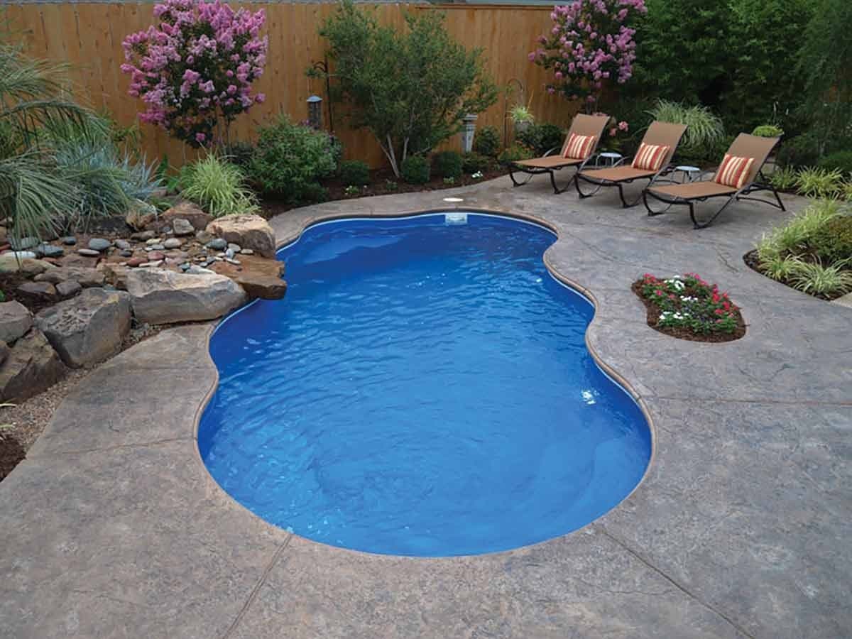 Liberty Victory fiberglass pool, freeform pool with a stamped concrete deck, boulders, flower beds and lounge chairs along a wood fence, manufacturer photo.