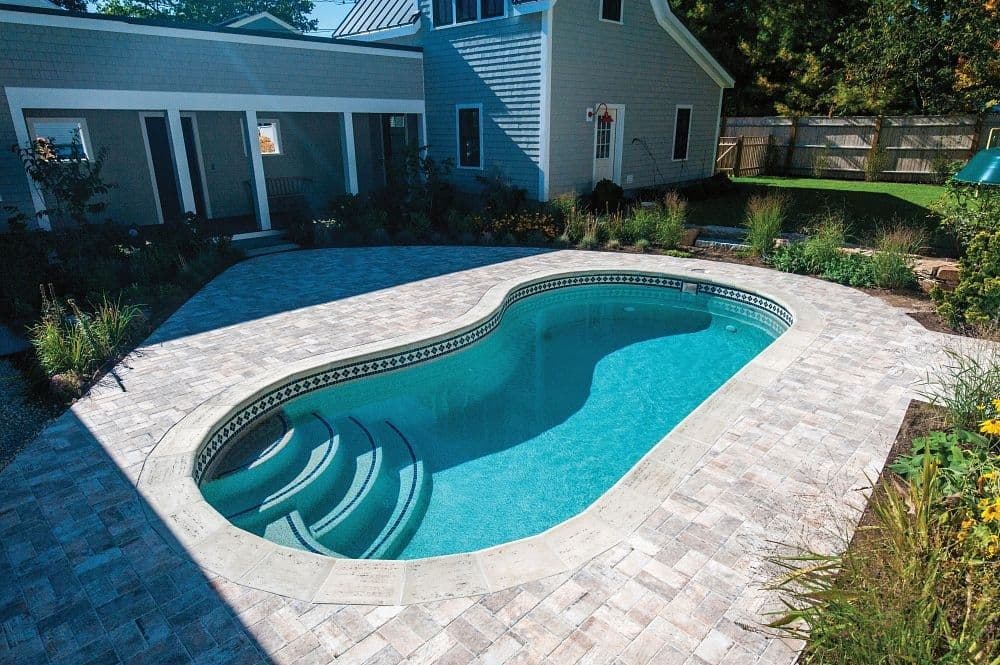 Liberty Virtue fiberglass pool, kidney-shaped pool with corner entry steps and a stone paver deck beside a blue house, seen from above, manufacturer photo.