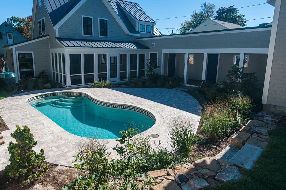 Liberty Virtue fiberglass pool, kidney-shaped pool with a stone paver deck, garden beds and a gray shingled house, manufacturer photo.
