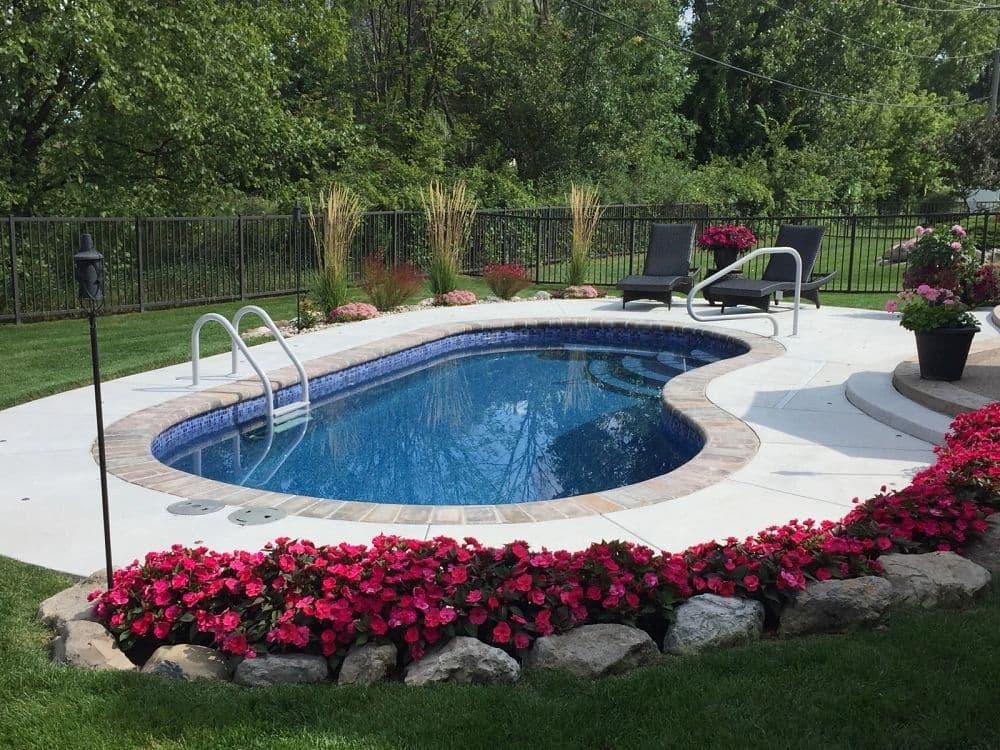 Liberty Virtue fiberglass pool, kidney-shaped pool with a handrail and red flowers along a light concrete deck, manufacturer photo.