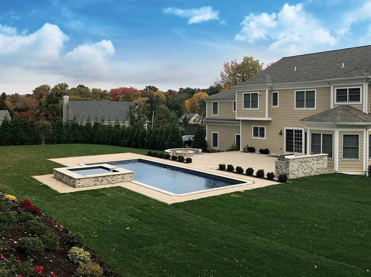 Liberty Virtuous fiberglass pool, long rectangular pool with an attached spa, a lawn and a large tan house behind, manufacturer photo.