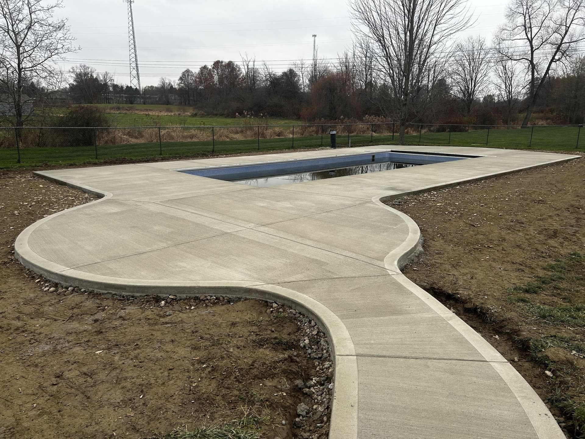 Rectangular pool surrounded by a newly poured concrete deck, with a wide curved concrete walkway leading away across bare soil toward a fenced field.