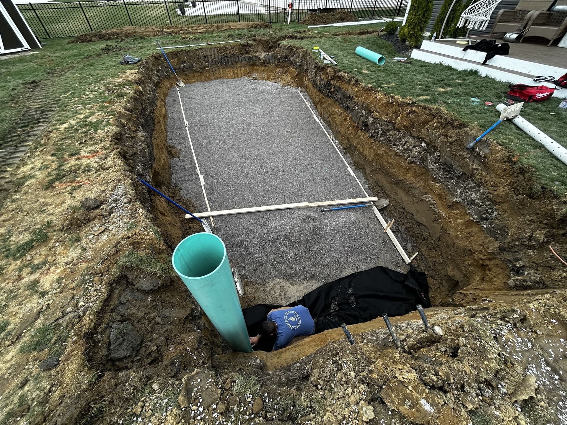 Rectangular pool excavation with a leveled gravel base marked by string lines, a vertical drain pipe in one corner and plumbing lines along the edge.