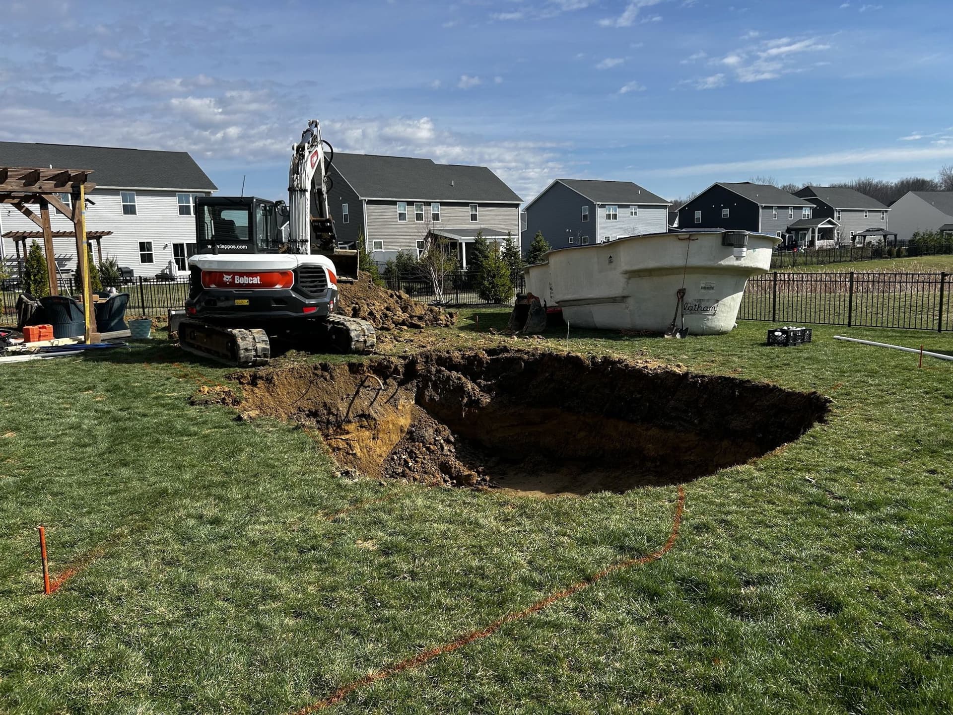 Freshly dug pool hole in a suburban backyard lawn with a compact excavator beside it and a white fiberglass pool shell resting on the grass nearby.