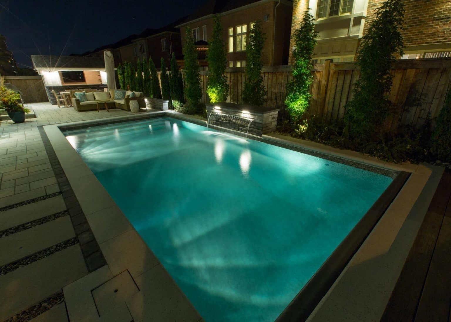 Rectangular pool lit with underwater lights at night, with a stone deck, landscape lighting and an outdoor lounge area behind it.