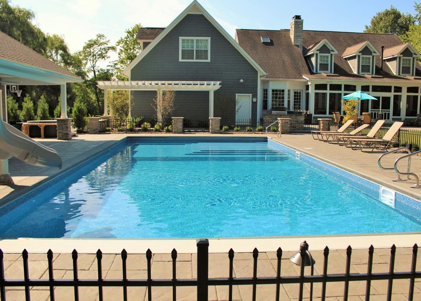 Large rectangular pool with a slide and lounge chairs in front of a gray shingle-style house, seen through a black metal fence.