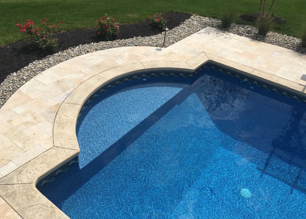 Vinyl liner pool with a rounded Roman-end step area and a patterned blue liner, framed by a tan paver deck and gravel border.